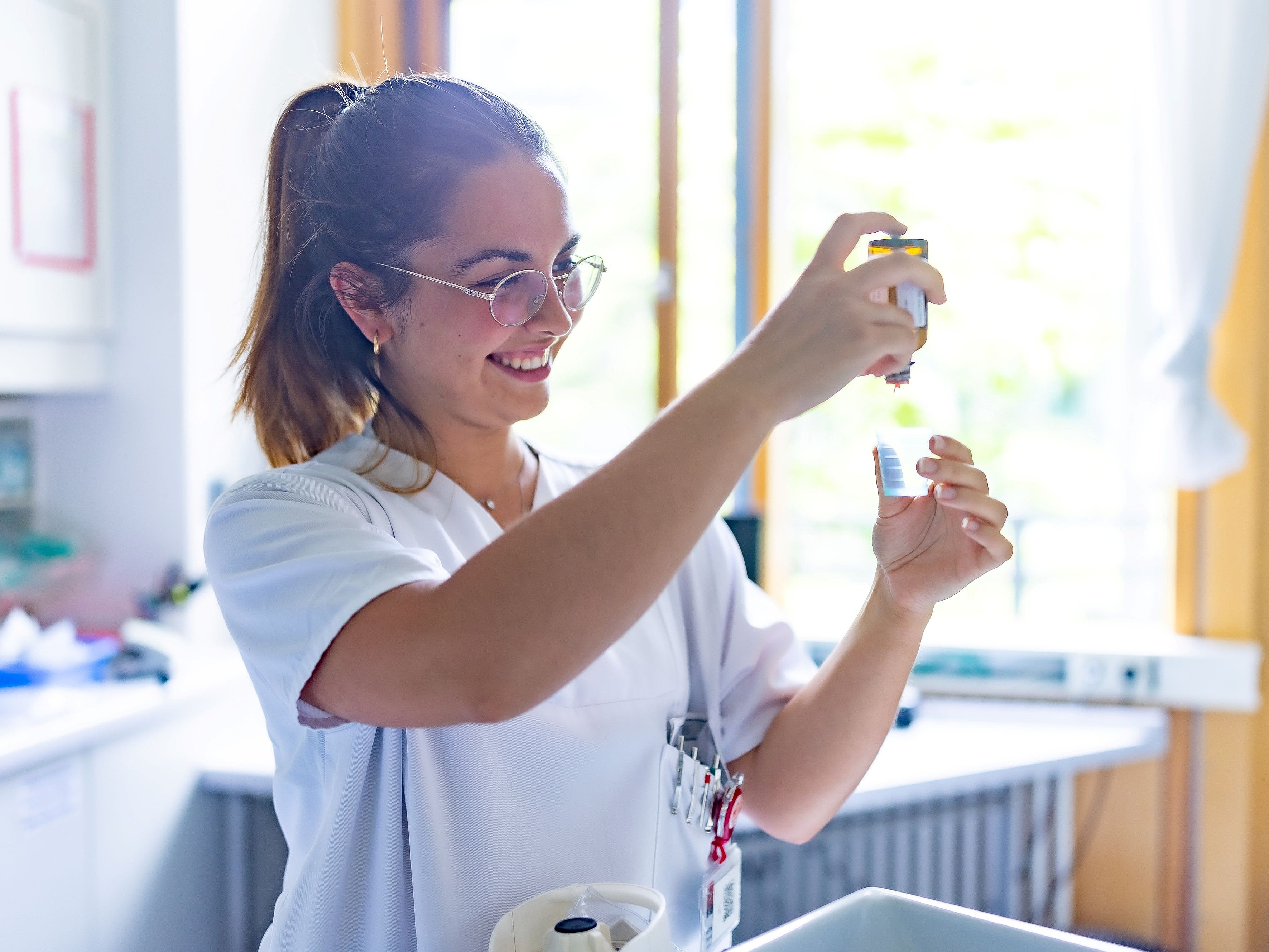Eine lächelnde junge Frau in einem weißen Kasack mit Brille hält konzentriert zwei kleine Behälter in den Händen, zur Vorbereitung von Medikamenten.Eine lächelnde junge Frau in einem weißen Kasack mit Brille hält konzentriert zwei kleine Behälter in den Händen, zur Vorbereitung von Medikamenten.