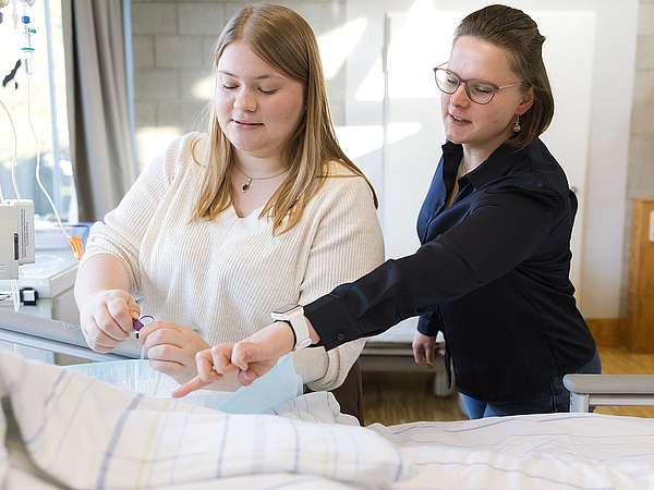 Eine Lehrerin der Berufsfachschule Agatharied gibt einer Schülerin Anweisungen an einem Pflegebett, während die Schülerin eine praktische Übung durchführt.