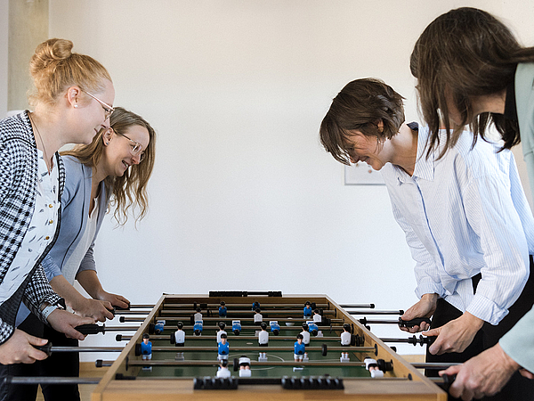  Vier Frauen spielen begeistert zusammen Tischkicker in einem Pausenraum.