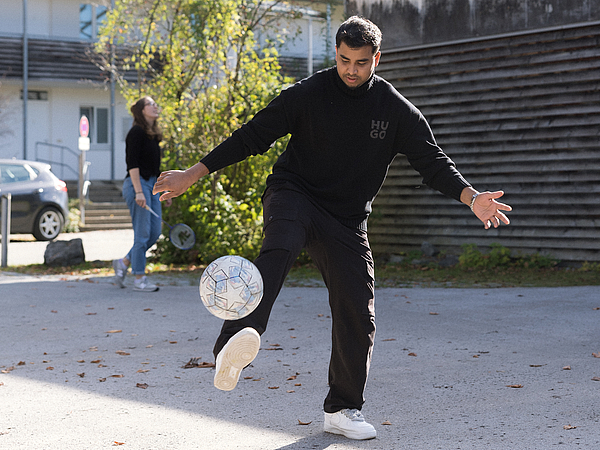 Ein junger Mann auf dem Gelände der Berufsfachschule Agatharied kickt einen Fußball mit dem Fuß nach oben. Im Hintergrund spielt eine andere Person Federball.