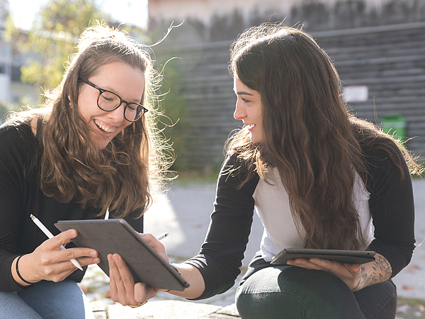Zwei junge Frauen, Auszubildende der Berufsfachschule Agatharied, sitzen im Freien und schauen lachend zusammen auf ein Tablet, das sie zum Lernen nutzen.