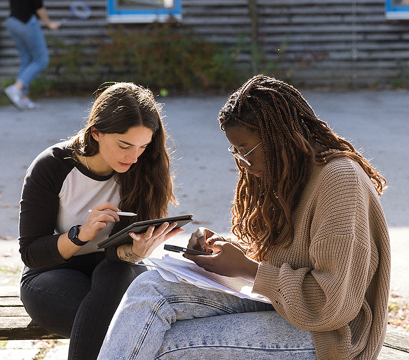 Austausch und digitales Lernen unter Auszubildenden Zwei junge Frauen sitzen im Vordergrund im Freien und nutzen digitale Geräte zum Lernen. Im Hintergrund ist ein Mann zu sehen, der einen Fußball mit dem Fuß nach oben kickt.