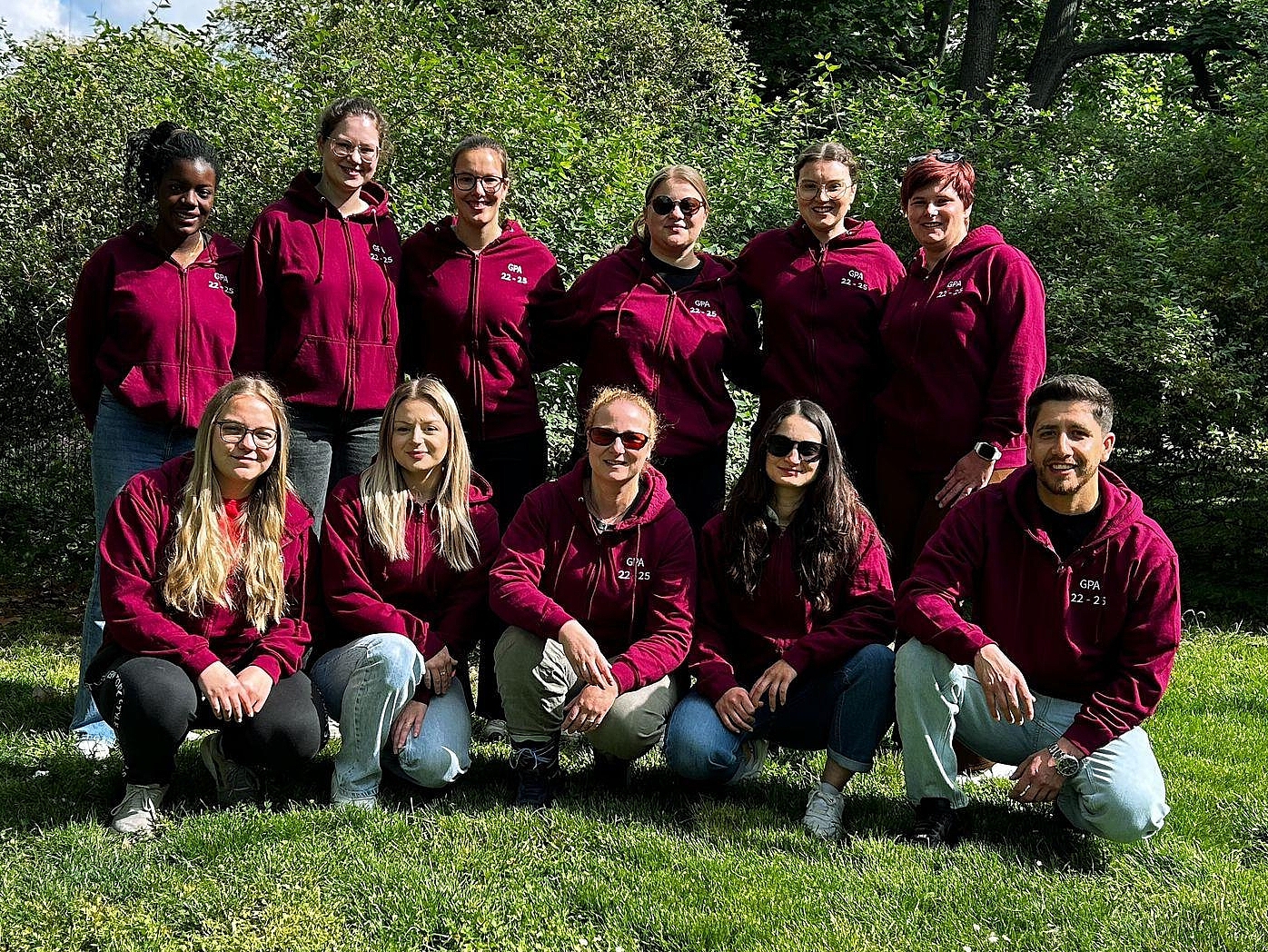 Gruppenfoto von elf Auszubildenden der Berufsfachschule Agatharied, die in bordeauxroten Hoodies in einem Park lächelnd in die Kamera schauen. Einige stehen, andere hocken im Vordergrund.Gruppenfoto von elf Auszubildenden der Berufsfachschule Agatharied, die in bordeauxroten Hoodies in einem Park lächelnd in die Kamera schauen. Einige stehen, andere hocken im Vordergrund.